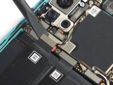Используйте плоский конец лопатки, чтобы отсоединить два шлейфа, закрывающих батарею смартфона OnePlus 8T
